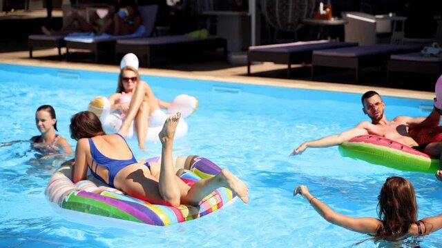 Friends chilling in private villa swimming pool, lie in the sun on inflatable flamingo, swan, floaties. Young people relax on party at luxury resort on sunny day. Bikini girls sunbathing. Slow motion.