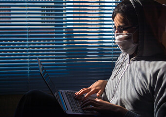 Young man with grey hoodie, face mask and glasses working/studying with a gray laptop on his legs. Sitting near a window with blue blinds,
illuminated with sunlight.