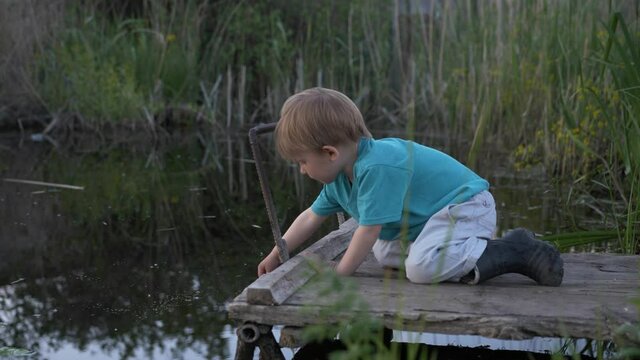 baby, little cheerful boy in rubber boots loves to play on pier in river while relaxing in village background of reeds during rural weekend