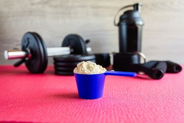 Plastic spoon or measuring scoop of whey protein on the background of sports equipment, a shaker for mixing the mixture, black dumbbells, leg ties, elastic bands for traction and attachment handles.