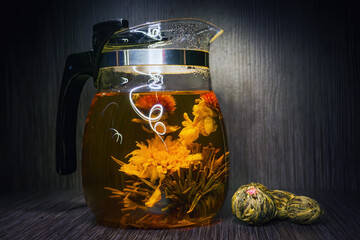 Blooming tea flower in glass teapot and dry flowering tea balls in dark indoors.