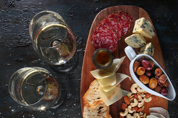 Two glasses of white wine and a selection of snacks on a dark background. Cheese and cold cuts with nuts on a wooden Board. Textured aged dark background. Copy of the space. Top view.