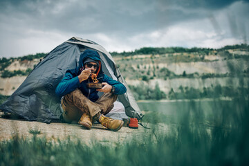 Young attractive man sitting by the tent and eating his lunch. Weekend camping concept.