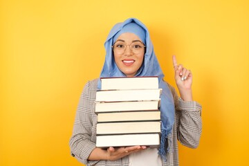 Young beautiful muslim woman wearing hijab and holding books on her arms against yellow background rising one finger up. Successful young student. 