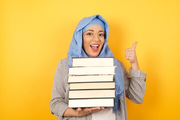 Young beautiful student woman wearing muslim hijab holding books doing happy thumbs up gesture with...
