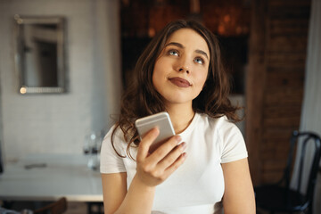 Technology and online communication concept. Portrait of happy thoughtful young woman in white top posing at home with mobile phone, looking up with pensive smile, messaging with boyfriend