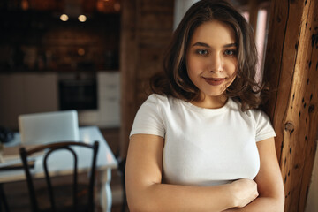 Fototapeta na wymiar Horizontal image of joyful cute young female freelancer in casual white top crossing arms on her chest having confident facial expression, working from home with open laptop in background