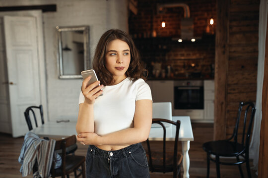 Charming cute young Caucasian female in casual clothes holding smart phone having thoughtful facial expression, posting pictures via social media account, thinking about caption to her photos