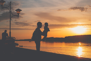 A sillhouete of mother and her baby playing together on the river side during sunset. Mom holding her toddler in the air and showing amazing view.