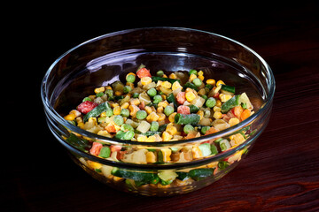 Frozen different vegetables in a glass plate on a wooden background.