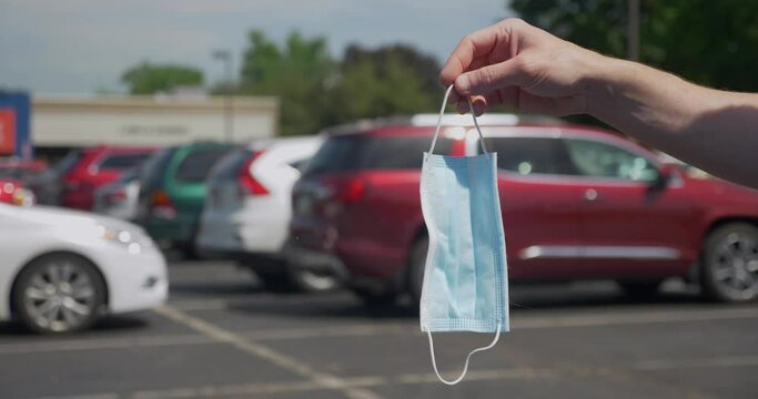 A man holds up then drops a used surgical face mask in a store's parking lot. Littering concept. Shot at 48 slow motion.  	