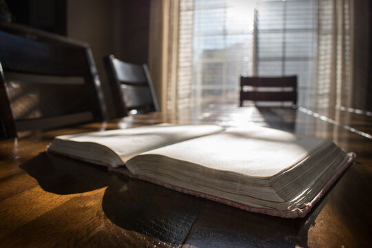 An open vintage book sits on a wooden table illuminated by sunlight from a nearby window.