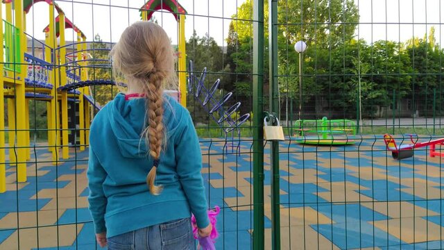 Sad lonely child near the closed playground because of the quarantine.A girl with long blonde braided hair staying alone near empty fenced showground without children.COVID-19 lockdown social distance