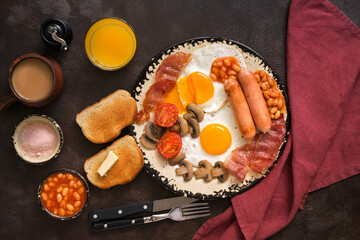 English traditional breakfast on a dark brown rustic background. Top view, flat lay. Fried eggs with bacon, sausages and beans, coffee, orange juice