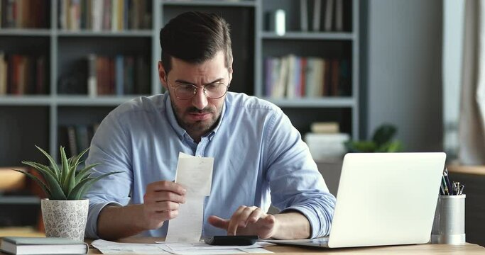 Frustrated young man in eyewear holding paper bill, calculating monthly expenses in online banking application on computer. Stressed businessman using software, managing financial data, money problem.