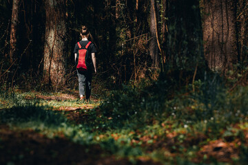 Lonely young girl with backpack goes along picturesque path into dark forest. Female teenager wandering in woods or lost. Free life of youth, independence or freedom of choice. Run away from home