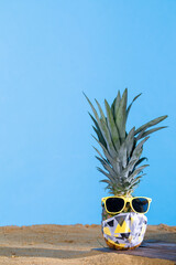 Pineapple in sun glasses and a protective mask stands on the sandy sea beach.
