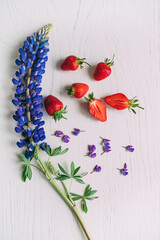A branch of lupins and strawberries. Still life from a serene branch of lupins and a few strawberry berries whole and cut on a white wooden  table, flat lay, vertical frame