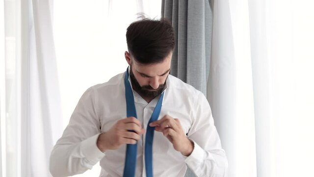 Young bearded handsome man in white shirt trying to tie his neck tie preparing for first job interview or date and having issue because it is his first time and it is complicate