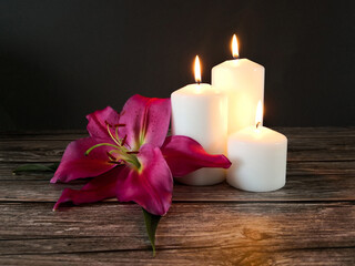 Close up of three pillar burning candles and pink lily flower on dark wooden table with black background. Memorial and remembrance concept.