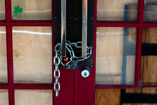 A Red Door With Partially Boarded Up Glass Windows And A Shamrock Sticker, Locked With A Metal Chain And A Padlock During The Shelter In Place Order.