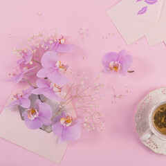 Delicate flatlay composition with morning cup of tea, pink letter bag full of purple orchid flowers and empty envelop on light pink background