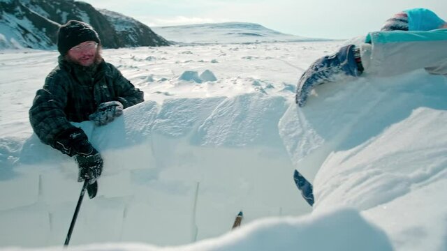 bearded man and woman in winter clothes build igloo with large snow bricks on frozen sea surface against brown hills