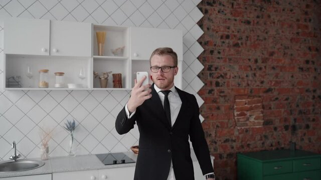 Young bearded businessman is having video call using phone while standing in home kitchen. American man talking about business and holding smartphone in hand, working at home during covid quarantine