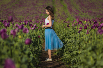 Obraz na płótnie Canvas Happy beautiful woman model in a blue skirt standing on a path of the blooming summer field full of purple poppy flowers 