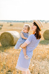 Pretty young mother in straw hat and striped dress holding on hands her cute little baby girl, looking away on the sky, walking in summer field with wheat hay bales on the background