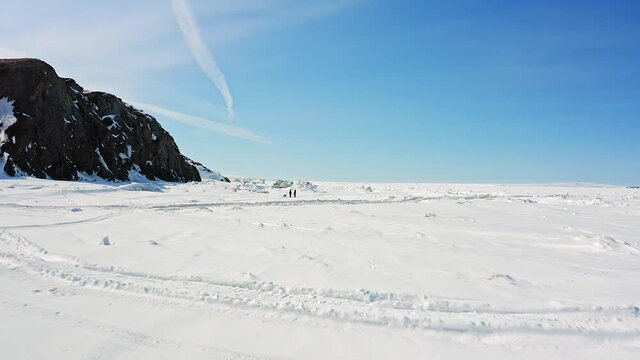 local people and tourist silhouettes wander on frozen sea surface near large brown hills on sunny winter day aerial motion