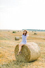 Naklejka na ściany i meble Young brunette Caucasian girl wearing straw hat and blue striped dress, sitting on a hay bale and posing at camera. Summer portrait of young woman in beautiful wheat harvesting field