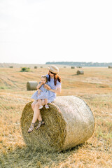 Naklejka na ściany i meble Happy family, mother and little daughter sitting on hay stack or bale in yellow wheat field in sunny summer day. Mommy and cute baby girl in striped dresses having fun during summer harvest time