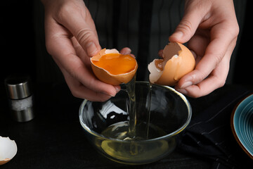 Woman separating egg yolk from white over glass bowl at black table, closeup - obrazy, fototapety, plakaty