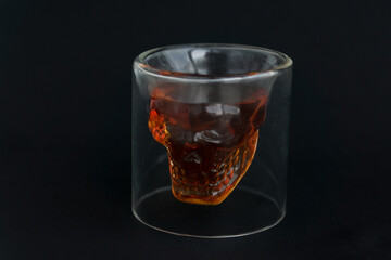 Close up low key image of skull shaped glass of brown alcohol drink and white smoke on black background. Mysterious atmosphere.