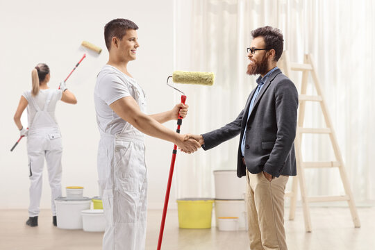 Male painter shaking hands with a bearded man and a female painter painting a wall inside a house