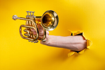 Hand showing a pocket trumpet through a torn hole in yellow paper background
