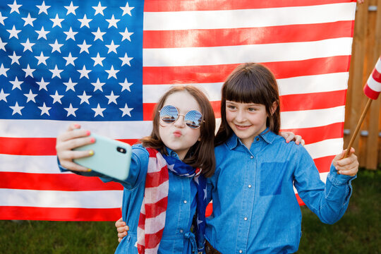 Two girls stand in the yard, holding the flag of USA and photographing themselves on a mobile phone