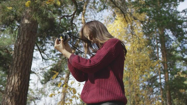 4K portrait of young brown-haired pretty female holding stillshot camera and taking photos of sunlit autumn forest. Low angle 360 degree tracking arc shot movement.
