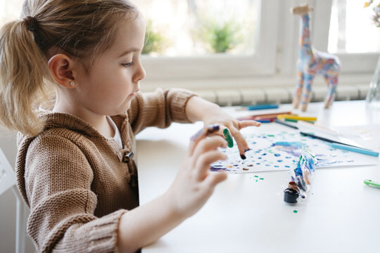 Cute child in casual outfit drawing paints with fingers while sitting at table with colour pencils and scissors at light kindergarten