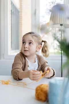 Little girl in casual wear winding woolen threads onto cardboard template to make handmade pompom for hat in cozy living room