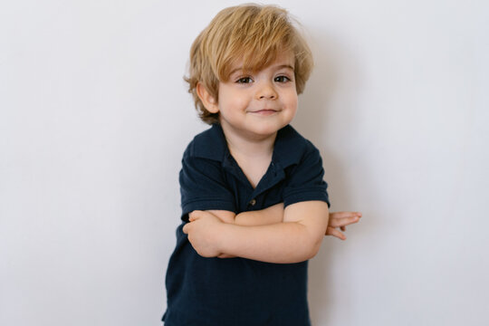 Adorable preschool boy in casual tee shirt with arms crossed smiling looking at camera leaning in a white wall background