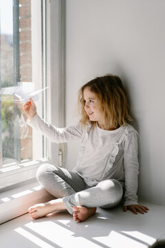 Happy blonde little girl in pajamas playing with paper airplane while sitting barefoot on window sill on sunny day