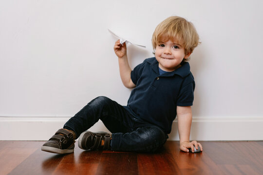 Cute blond boy in casual outfit sitting on wooden floor playing with paper plane smiling brightly at camera on white wall background