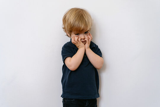 Annoyed blond little boy in casual clothes looking away with dissatisfaction while standing on white wall with hands on face