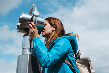 Anonymous blond female in blue coat looking through street coin operated binocular while standing on wooden pavement near concrete fence on sunny day