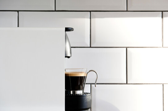 Modern pod coffee maker pouring hot espresso into glass cup on background of ceramic tile on kitchen wall