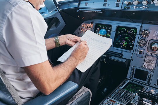 Side view of crop male aviator sitting in cockpit and filling in document while preparing for flight in modern aircraft