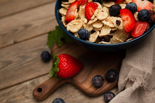 From above delicious breakfast bowl of corn flakes with strawberries and blueberries placed on cutting board and decorated with linen cloth and berries around dish on wooden background