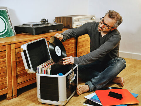 Stylish male wearing casual clothes and eyeglasses sitting on wooden floor and picking vinyl record while enjoying weekend at home